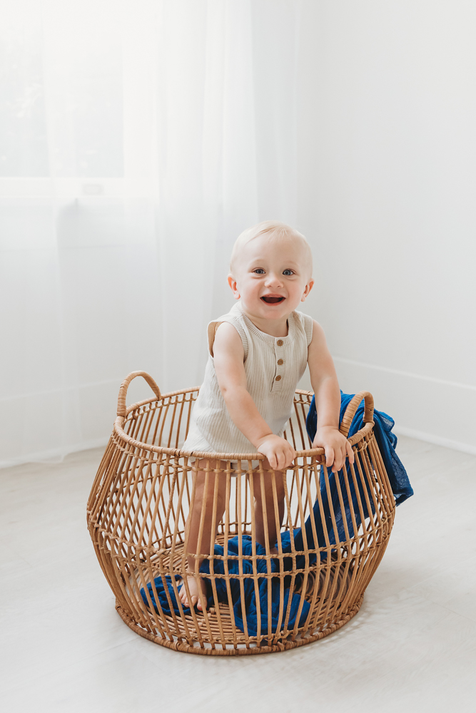 Smiling baby in cream ribbed romper sitting in wicker basket with blue fabric on white backdrop with soft natural lighting
