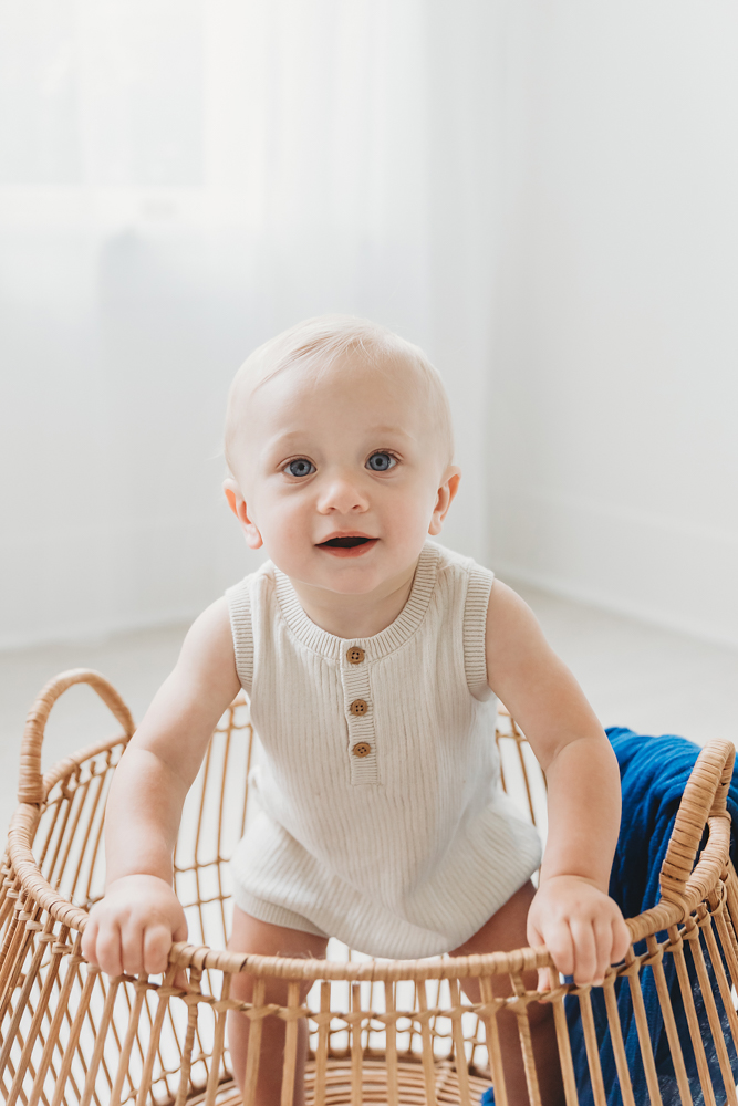 Baby in cream knit romper sitting in wicker basket on white backdrop, soft natural lighting by Tracy Rowell
