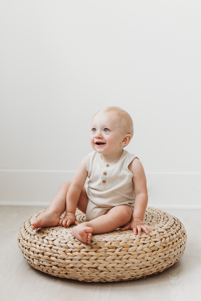 Smiling baby in cream knit romper sitting on woven basket against white backdrop - Reflection Images by Tracy Rowell