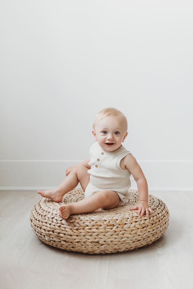 Smiling baby in cream knit romper sitting on woven basket cushion against white backdrop with soft natural lighting