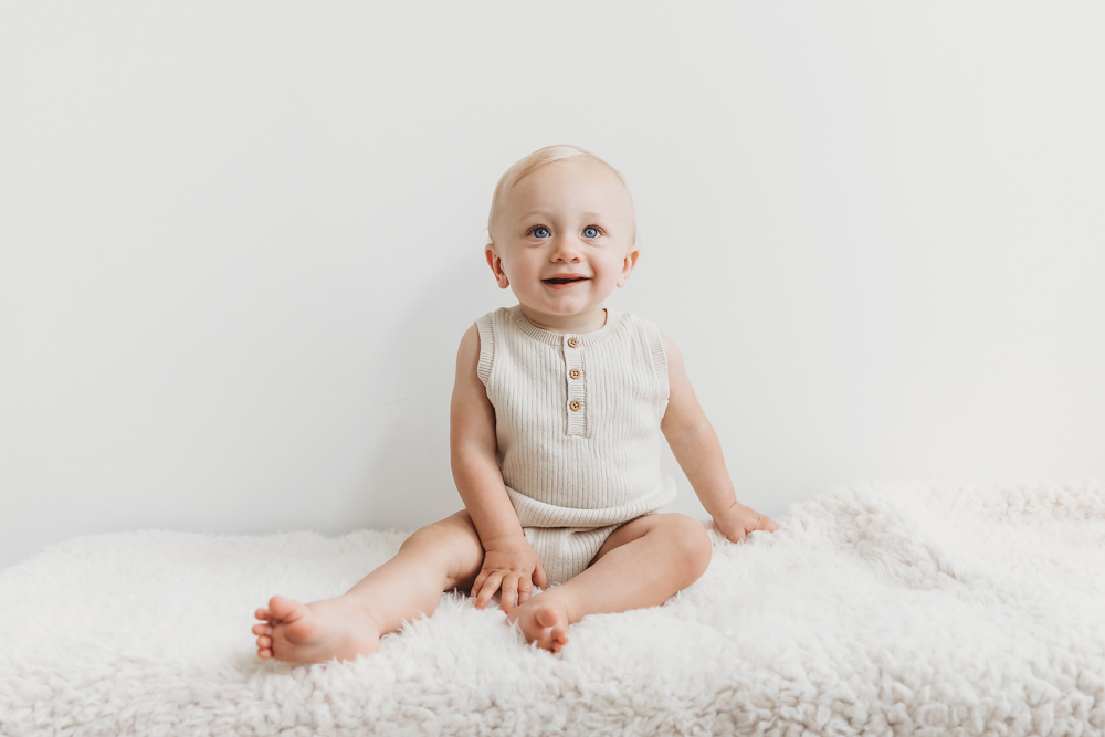 Happy baby in beige ribbed romper sitting on white fluffy rug against light gray backdrop, Tracy Rowell portrait