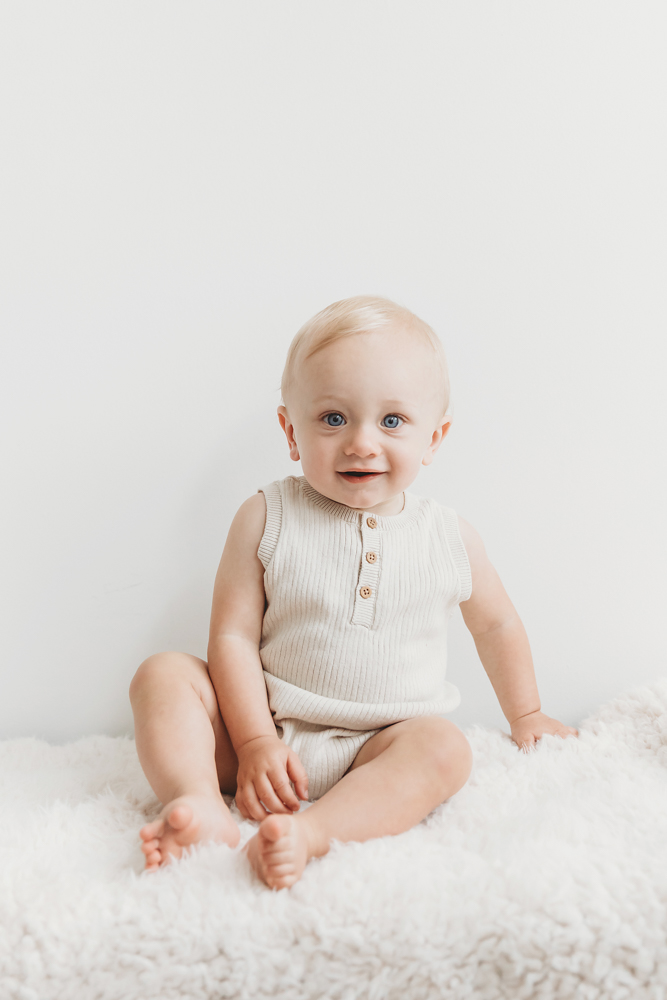 Smiling baby with blue eyes sitting on white fluffy blanket wearing cream ribbed romper against soft gray backdrop
