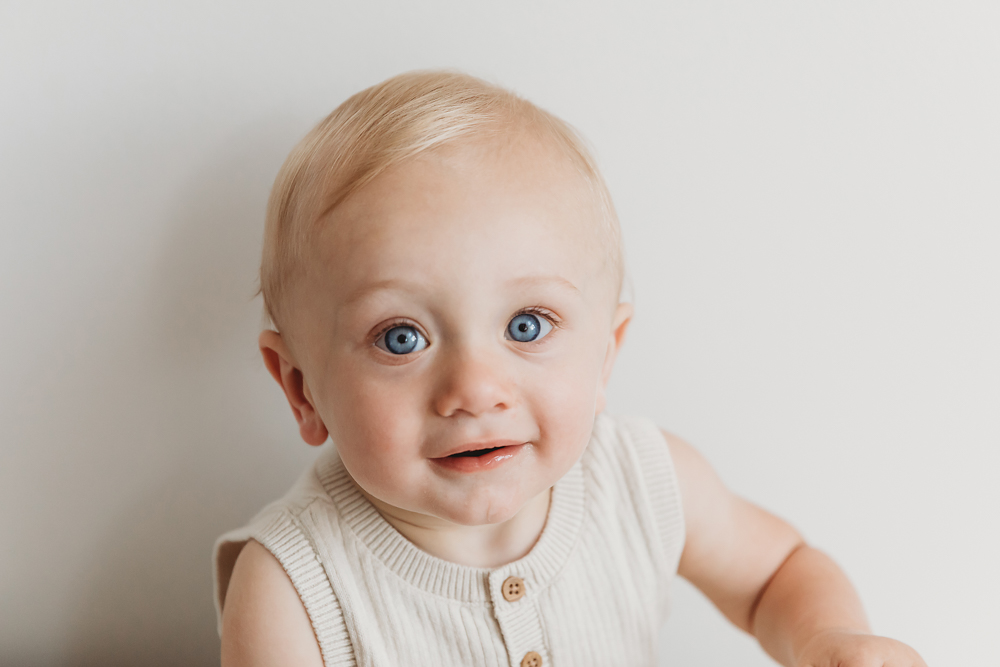Blue-eyed baby in cream knit vest smiling at camera against white backdrop, soft natural lighting, cake smash portrait