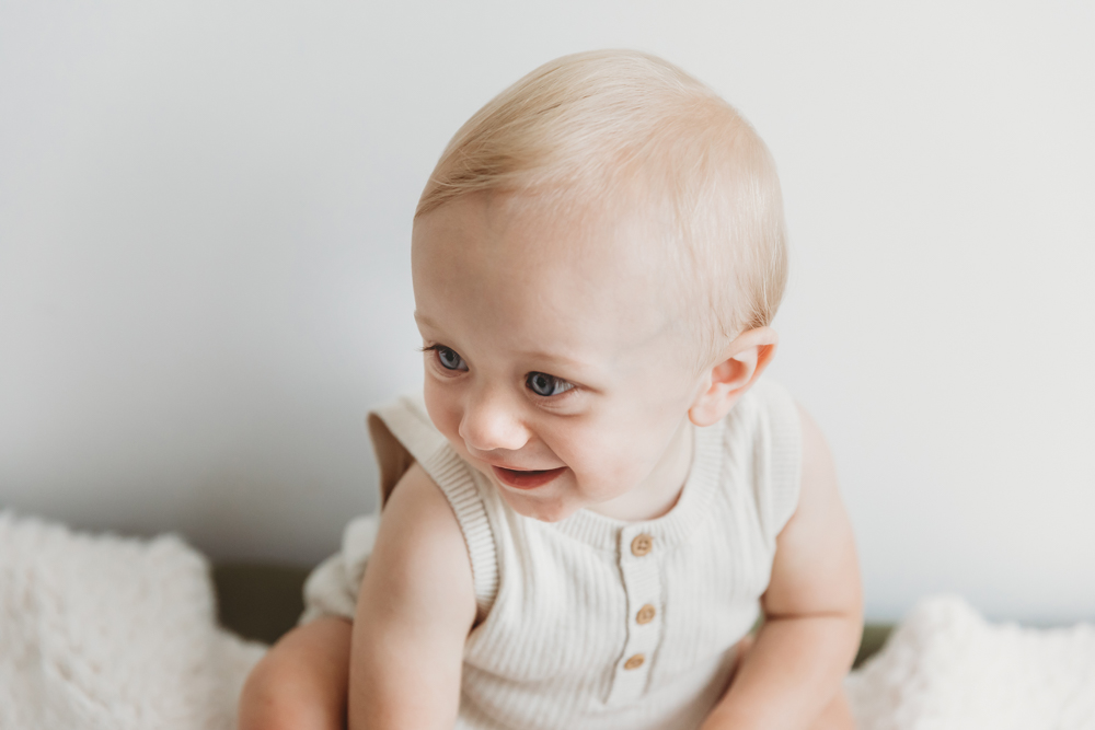 Happy baby with blonde hair in cream knit vest smiling at camera on white bedding with soft natural lighting