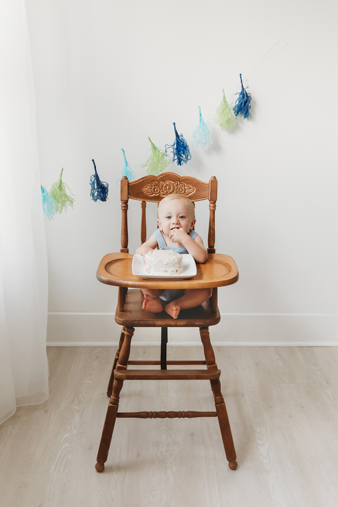Baby in wooden highchair eating cake during first birthday photoshoot with blue and green tassels on white wall