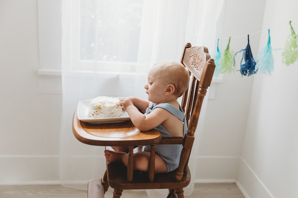 Baby in gray romper sitting in wooden high chair with white cake, colorful streamers hanging in bright room