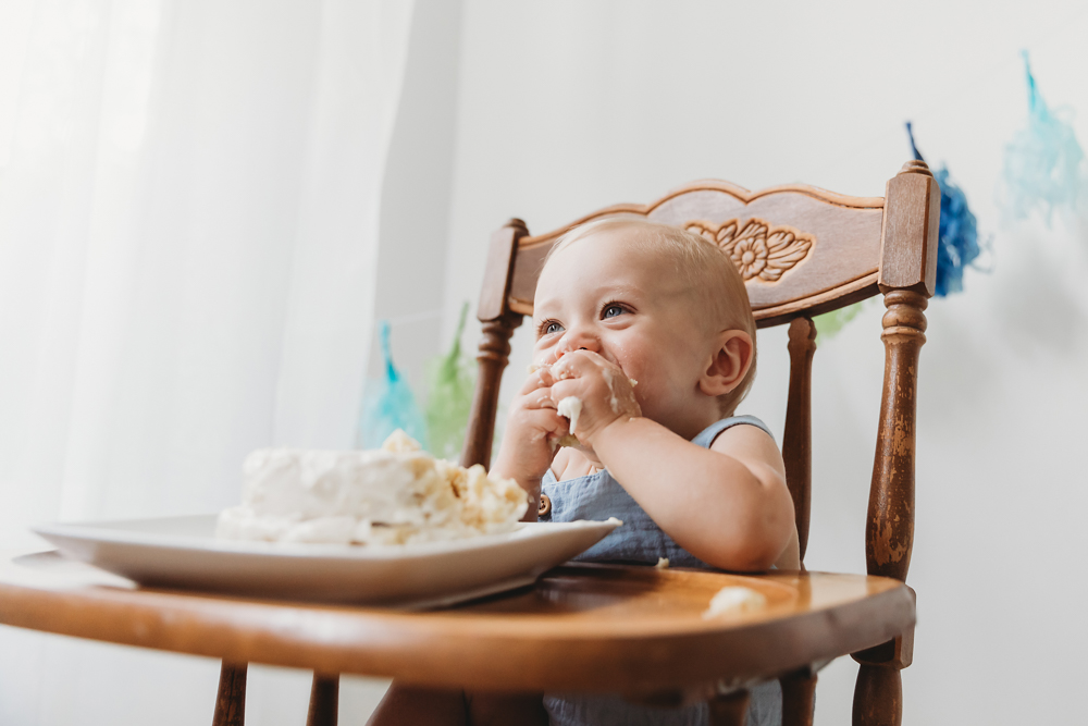 Baby in blue outfit sitting in wooden high chair eating cake, smiling with frosting on hands against white backdrop