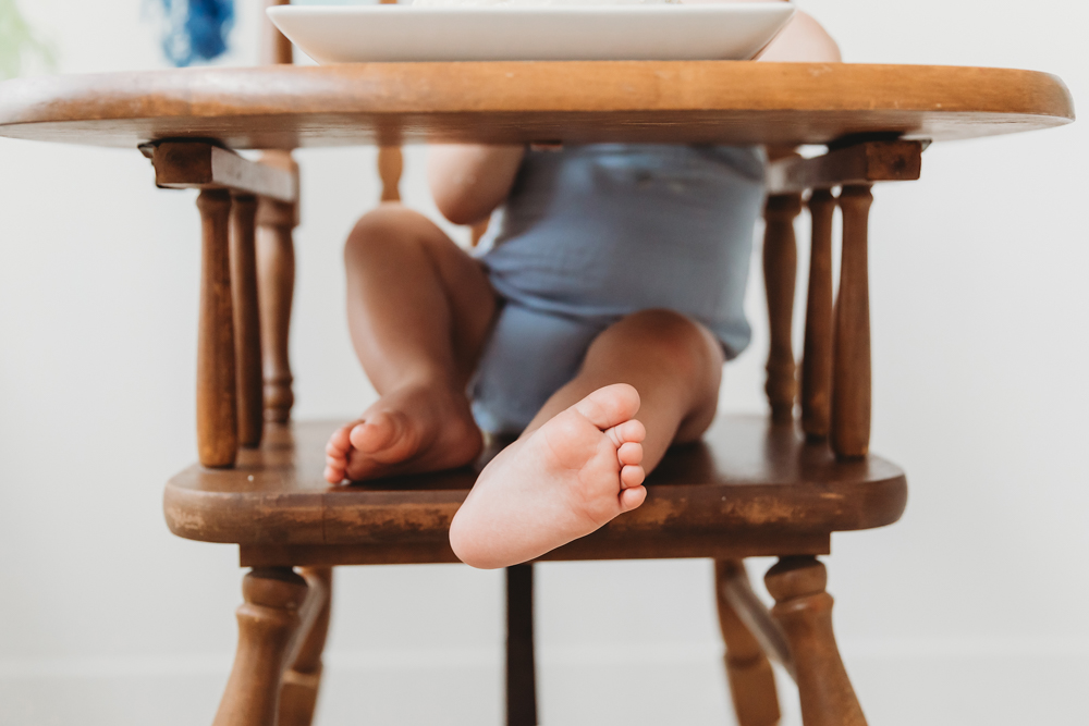 Baby in blue outfit sitting in vintage wooden high chair with feet dangling, soft natural lighting, professional photography studio