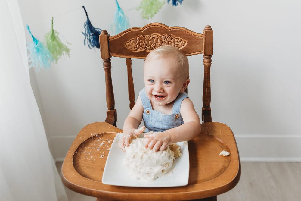 Smiling baby in light blue romper sitting in wooden high chair doing cake smash with white frosted cake