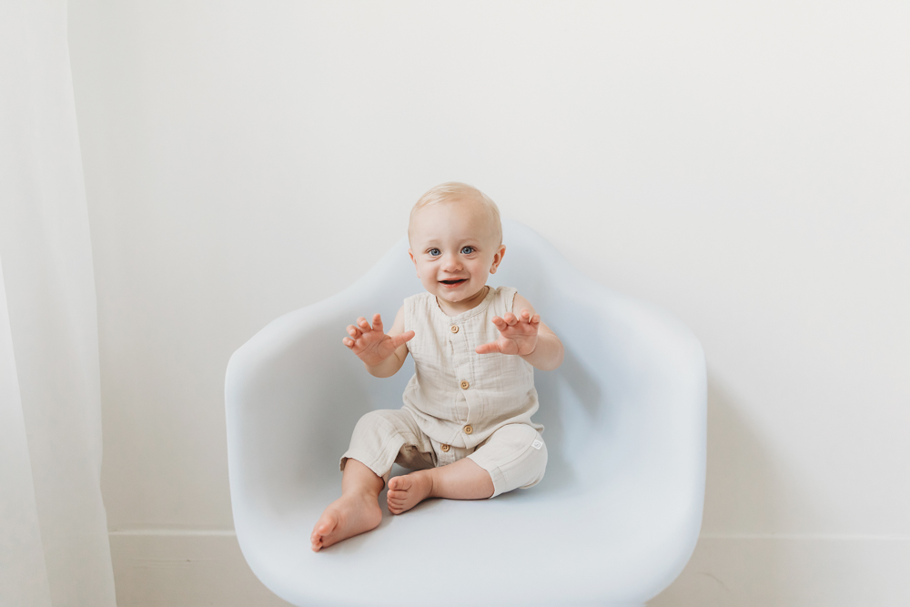 Smiling baby in cream linen romper sitting in white modern chair, hands raised playfully, soft studio lighting by Tracy Rowell