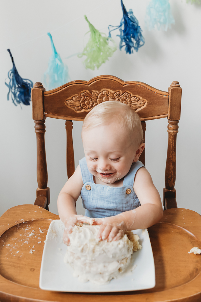 Smiling baby in blue overalls sits in carved wooden high chair enjoying messy cake smash session - Reflection Images by Tracy Rowell