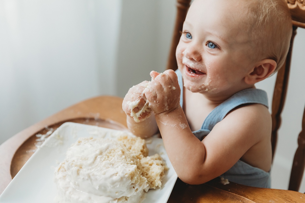 Smiling toddler in gray bib eating cake with hands in wooden high chair, face and hands covered in frosting