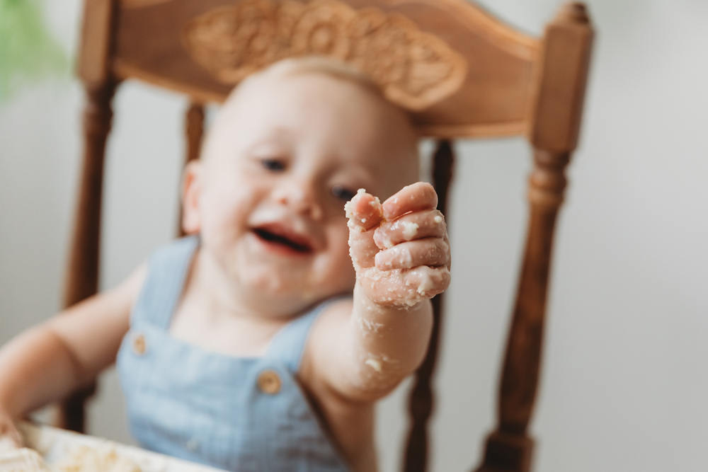 Happy baby in light blue outfit sitting in wooden high chair with messy hands from eating, Tracy Rowell portrait