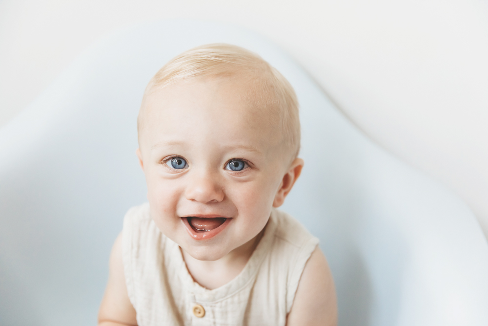 Smiling baby with blonde hair and blue eyes wearing cream sleeveless outfit against soft white backdrop