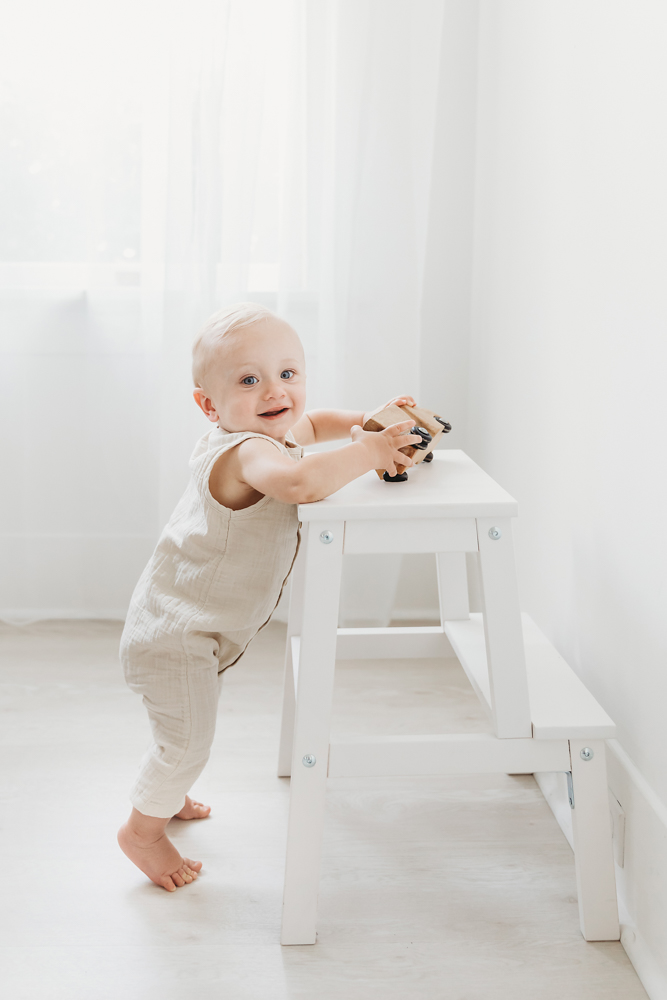 Smiling baby in beige romper stands by white wooden bench holding toy car, bright studio lighting for cake smash session