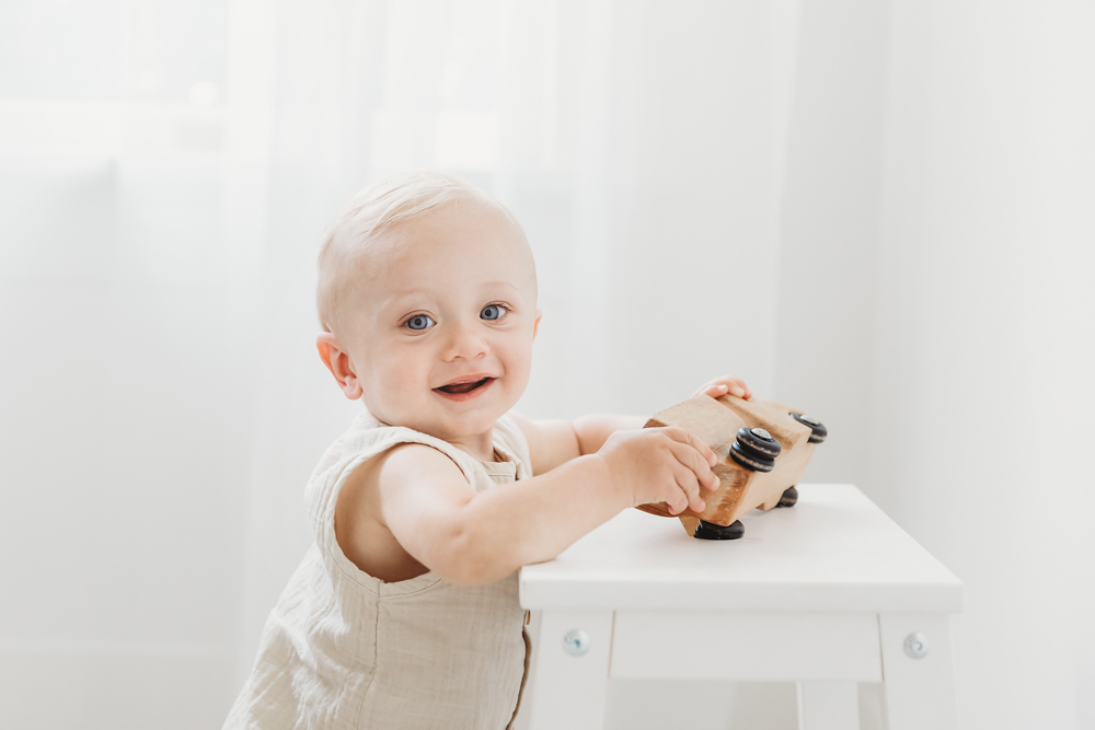 Happy baby in cream romper playing with wooden toy car on white cube against soft white backdrop