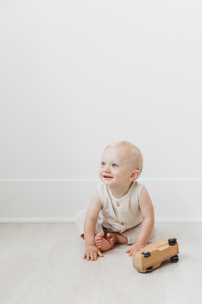 Smiling baby in cream linen romper sitting on white floor with wooden toy car during first birthday photoshoot