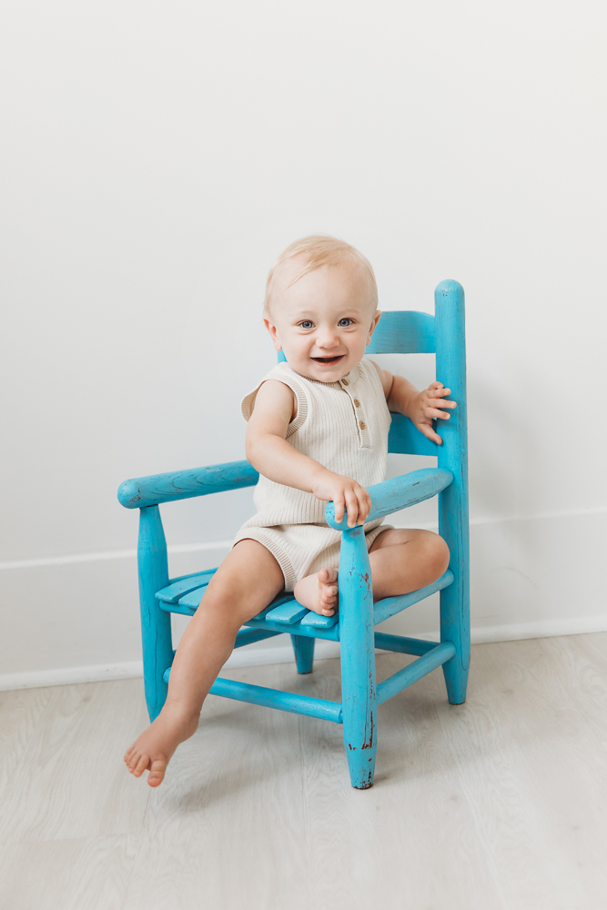 Smiling baby in cream romper sitting on vintage turquoise wooden chair against white backdrop with soft natural lighting