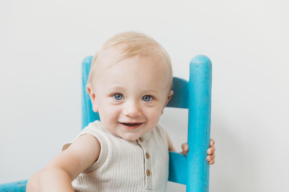 Smiling blonde toddler in cream ribbed tank top sitting on bright turquoise wooden chair, white backdrop, florence sc photographer