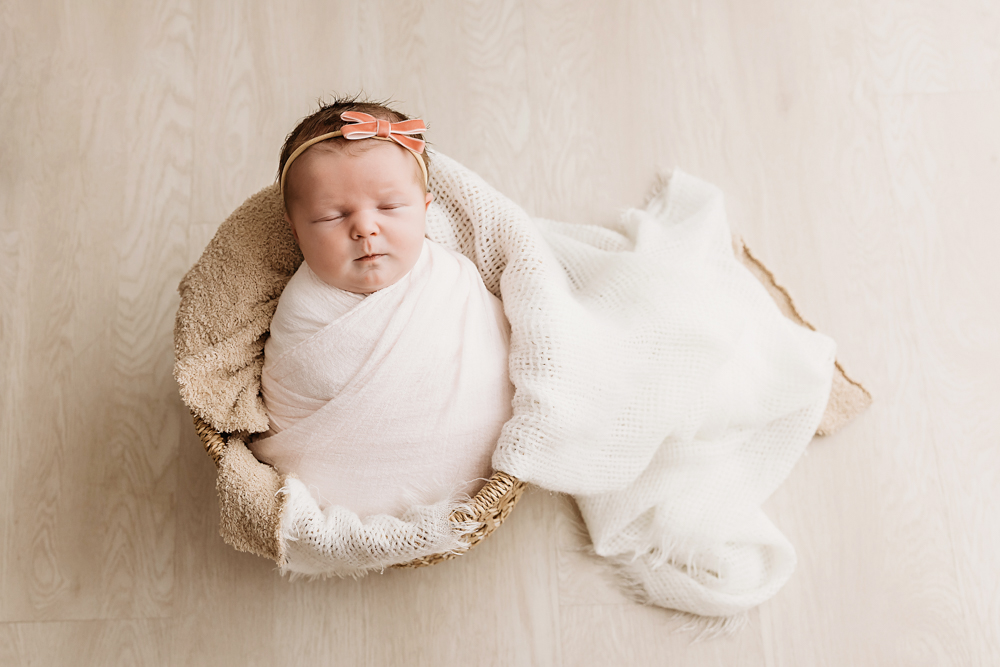 Sleeping baby in white wrap with coral bow headband nestled in wicker basket, professional newborn pictures on cream backdrop