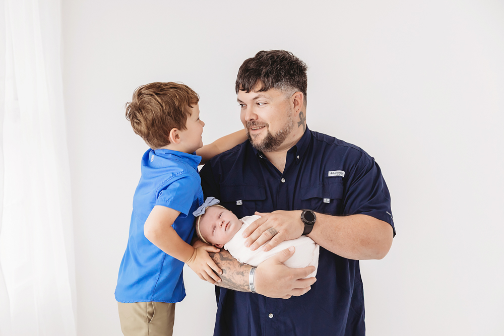 Father holding newborn baby while young son in blue polo stands beside them, white studio backdrop, professional photography studio near me