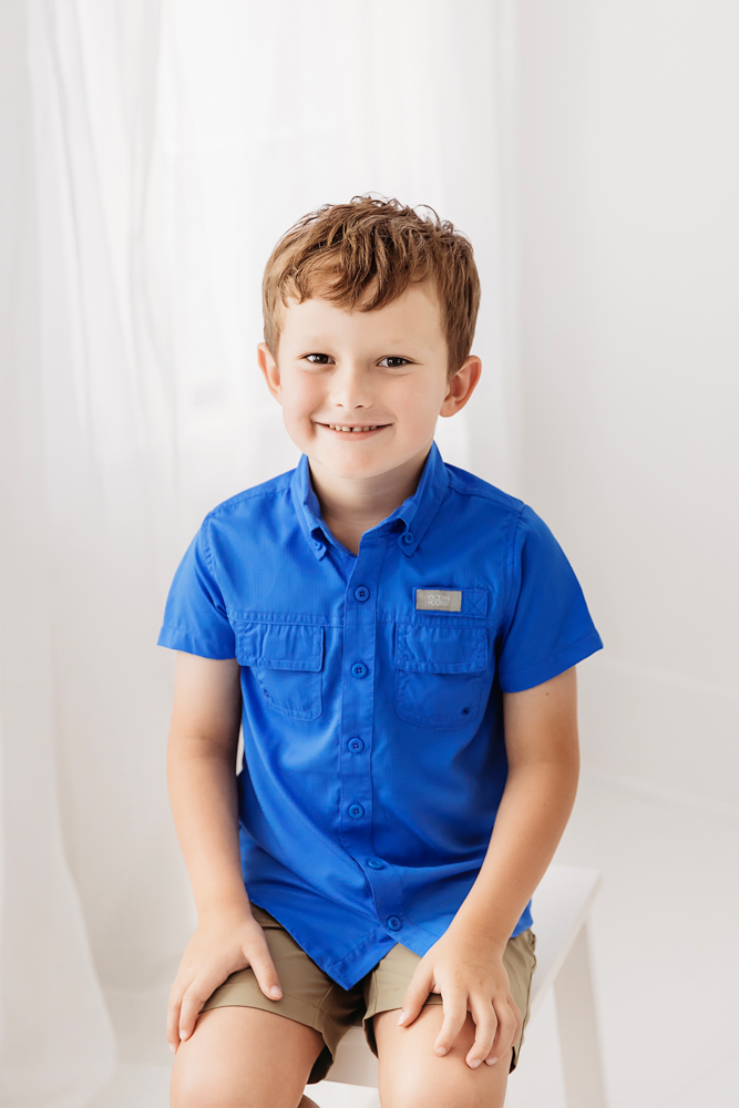 Smiling young boy in bright blue button-up shirt and khaki shorts against white backdrop, professional photographer near me