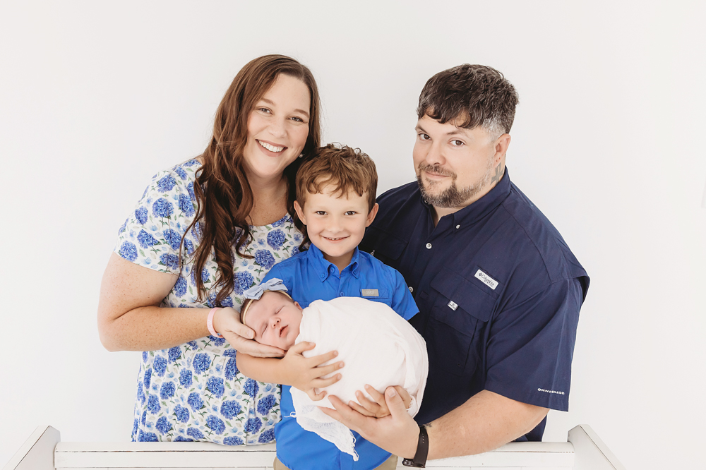 Family of four with newborn baby in professional photography studio - mother in blue floral top, father in navy shirt, child in blue, white backdrop