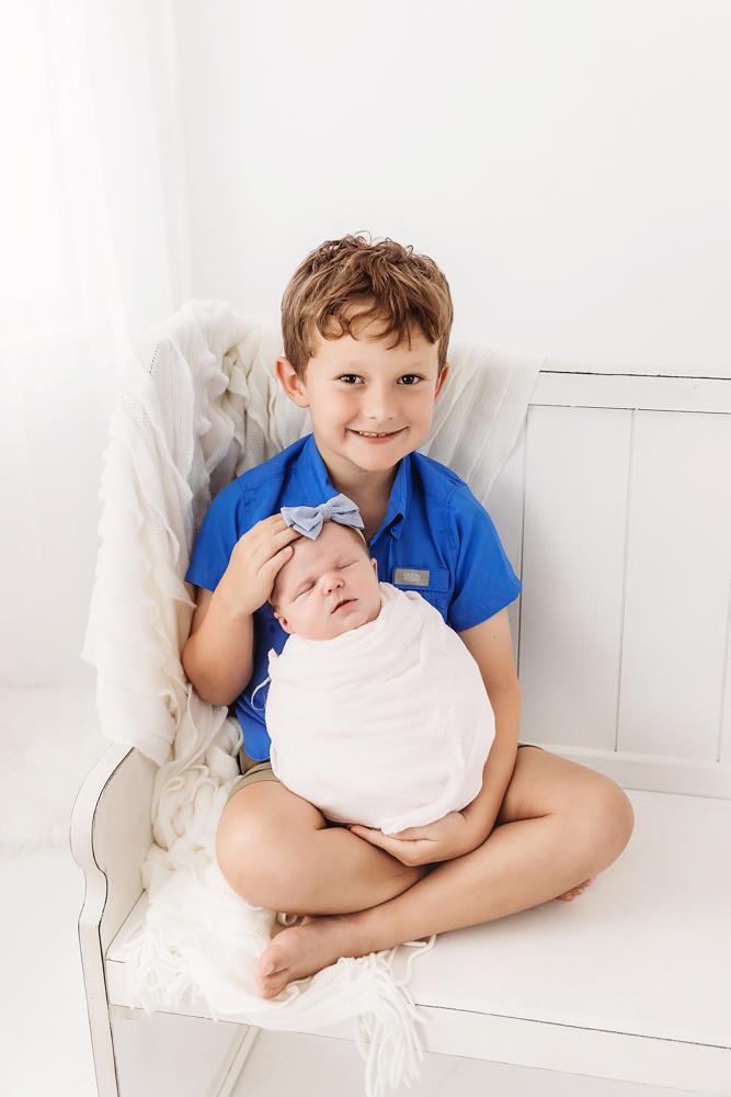 Smiling child in blue shirt holding swaddled newborn baby on white chair, soft lighting professional photography