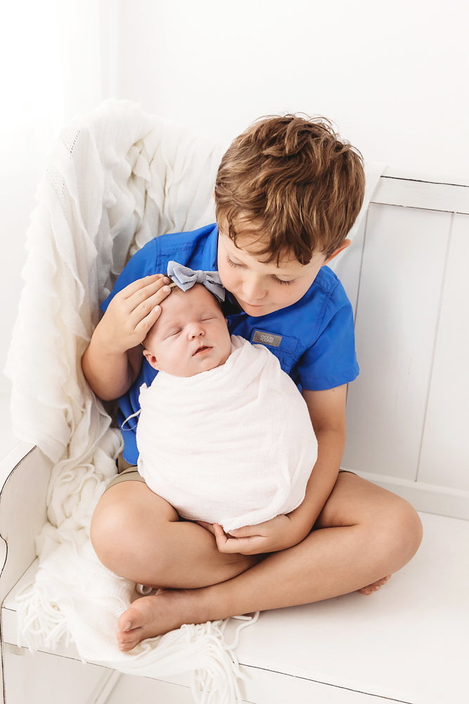 Boy in blue shirt tenderly holding newborn baby in white wrap on cream backdrop by professional photographer
