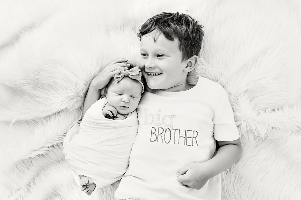 Smiling child holding swaddled newborn baby on white fur blanket, big brother shirt, black and white newborn photographer portrait