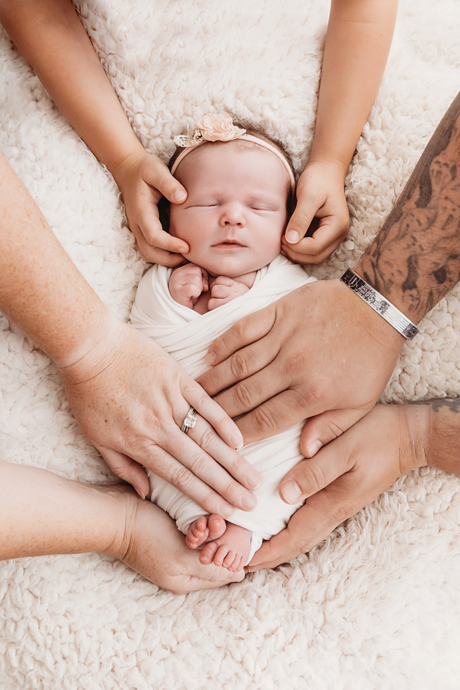 Newborn baby in cream wrap with pink headband surrounded by family hands on white textured blanket - Reflection Images by Tracy Rowell