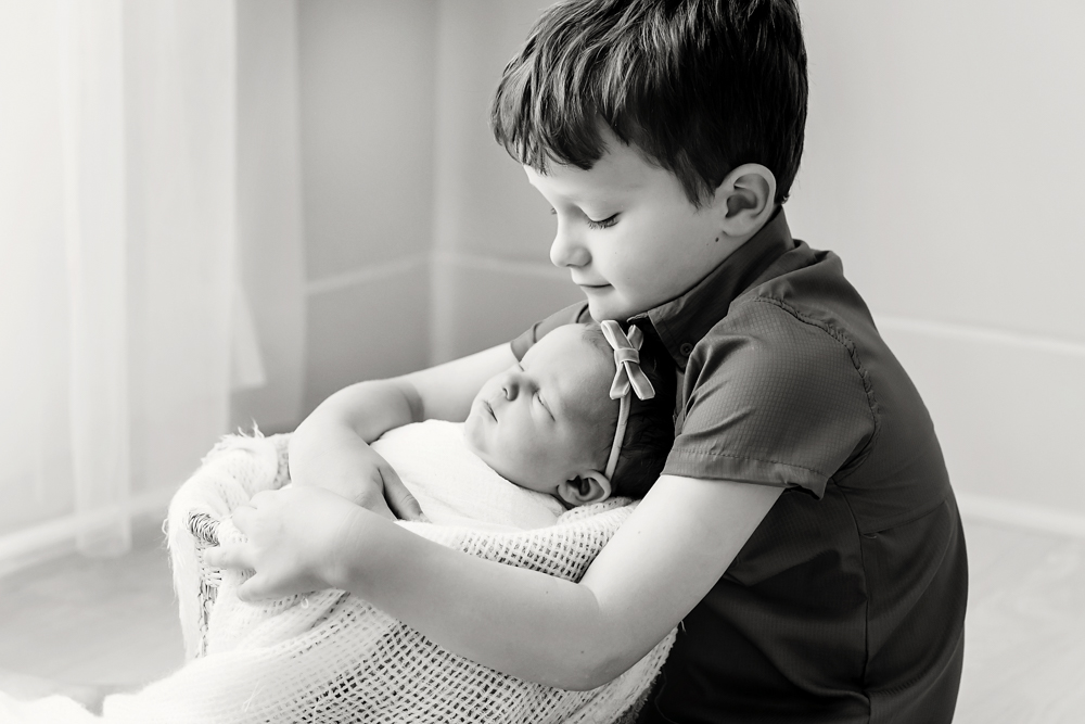 Young boy tenderly holding sleeping baby in white knit blanket, soft natural lighting, black and white newborn photography