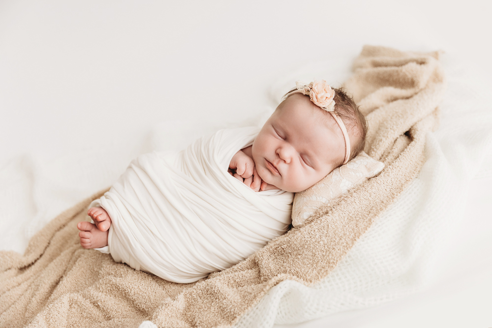 Sleeping newborn baby wrapped in cream striped swaddle with peach flower headband on beige textured blanket for newborn photos
