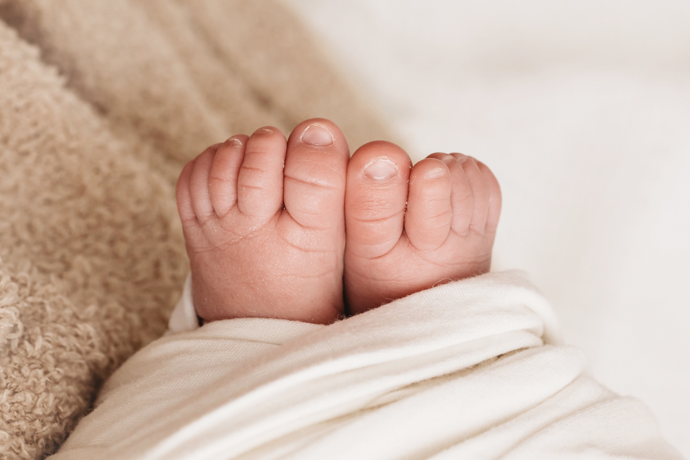 Tiny baby feet peeking from cream blanket on textured beige backdrop for soft newborn pictures portrait session