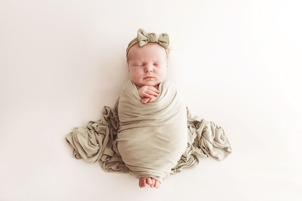 Peaceful newborn baby swaddled in sage green wrap with matching bow headband on cream backdrop, professional photography studio near me