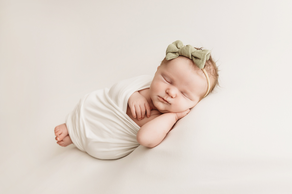Sleeping newborn baby in cream wrap with sage green bow headband on neutral backdrop by professional photographer near me