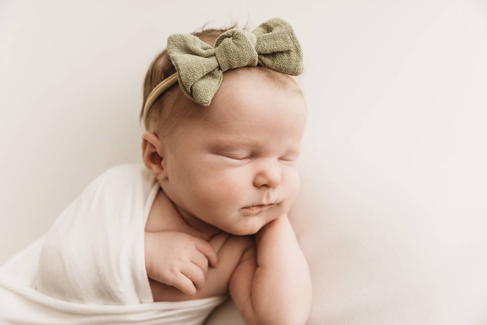 Sleeping newborn baby with sage green bow headband in white wrap on cream backdrop at professional photography studio