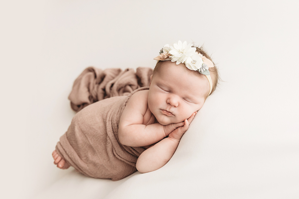Sleeping newborn baby in taupe wrap with white floral headband on cream backdrop, professional photography portrait