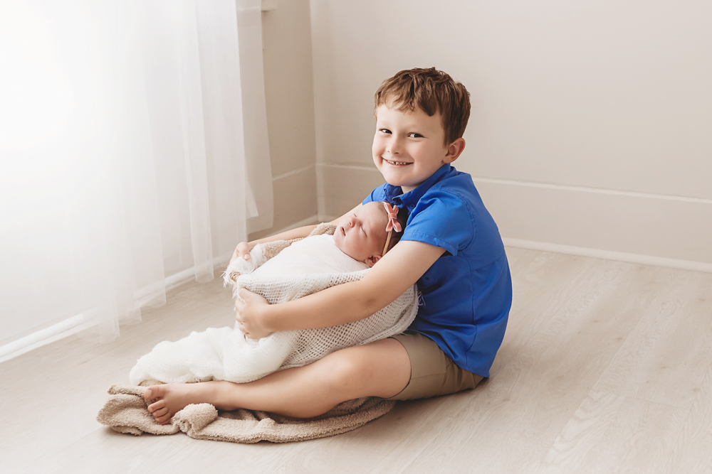 Smiling child in blue shirt holding swaddled newborn baby on cream blanket, soft natural lighting by newborn photographer