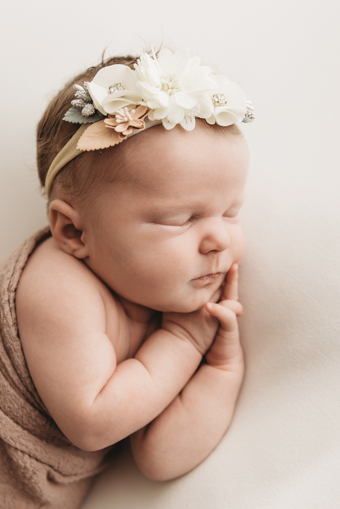 Sleeping newborn baby with white floral headband wrapped in brown knit blanket on cream backdrop by professional photographer