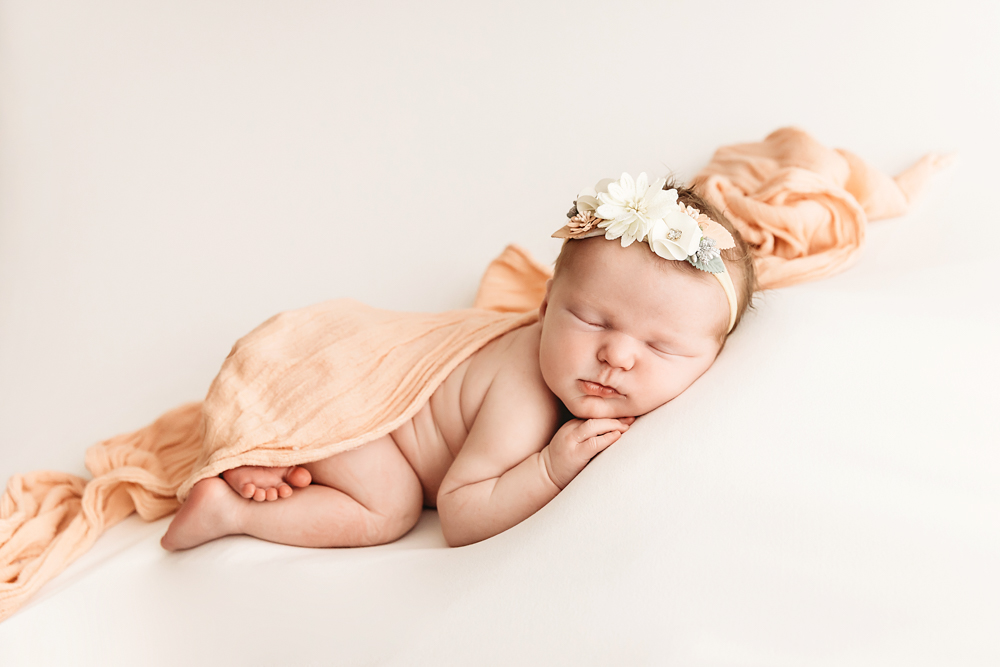 Sleeping newborn baby with white floral headband wrapped in peach fabric on cream backdrop for professional newborn pictures