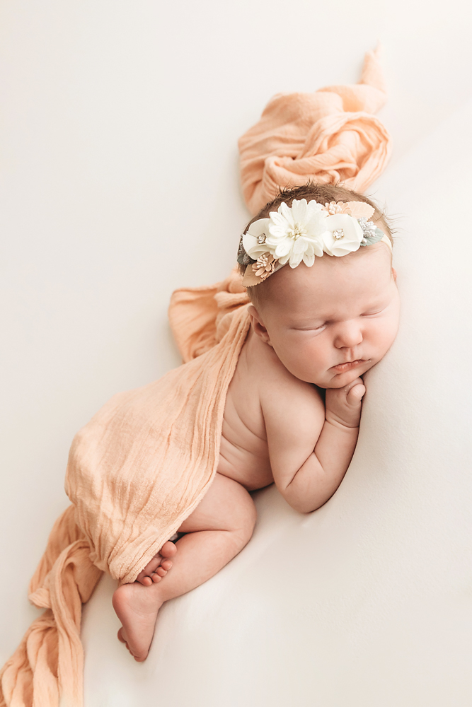 Sleeping newborn in peach wrap with white floral headband on cream backdrop, soft natural lighting newborn photography