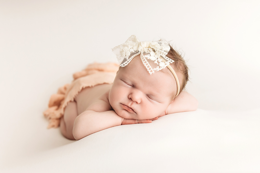 Sleeping newborn with white lace headband on cream backdrop, soft lighting - newborn photographer portrait session