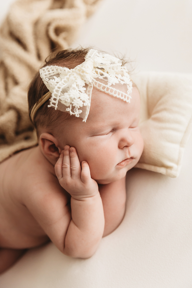 Sleeping newborn with white lace bow headband on cream backdrop, soft natural lighting by Tracy Rowell