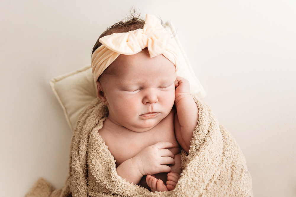 Sleeping newborn baby with peach floral headband wrapped in textured beige blanket on cream backdrop for newborn photos