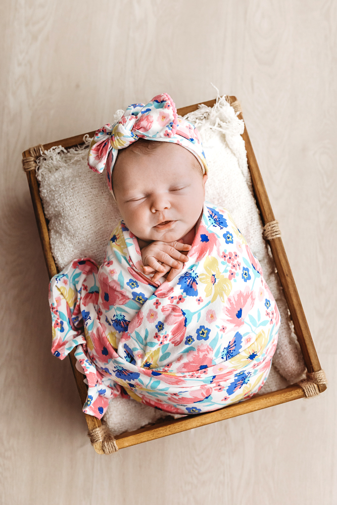Sleeping newborn in floral wrap and headband nestled in wooden crate for newborn pictures on cream backdrop
