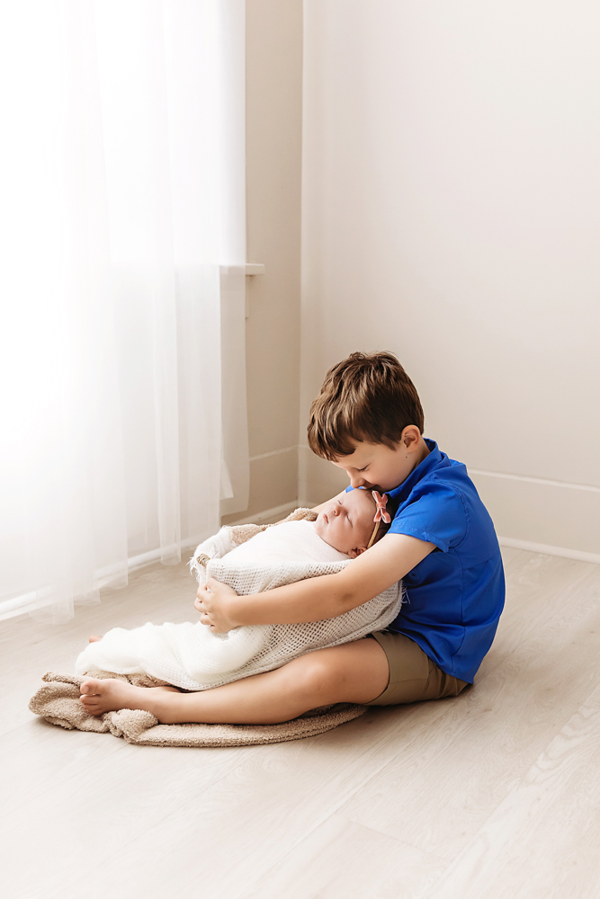 Young boy in blue shirt tenderly holding newborn baby in white blanket, sitting on beige rug in soft natural light studio
