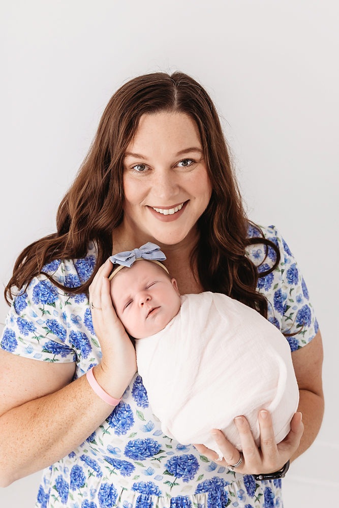 Smiling woman in blue floral dress holding sleeping baby in white wrap with blue bow headband for newborn photos