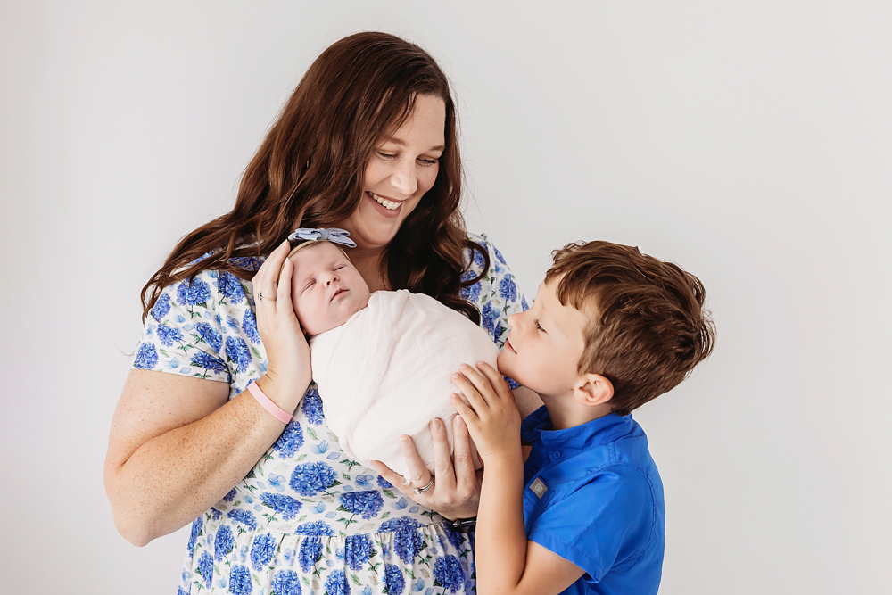 Mother in blue floral dress holds sleeping newborn in white blanket while young boy in blue shirt looks on, newborn pictures