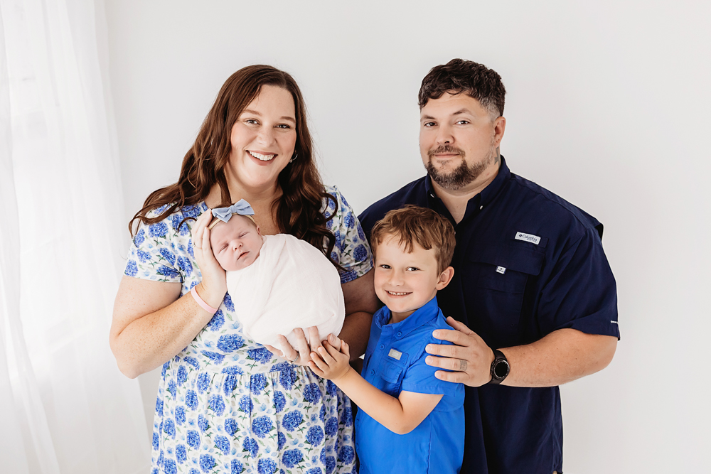 Smiling family of four with newborn baby in cream wrap, mother in blue floral dress, father and child in blue shirts, white backdrop by florence sc newborn photographer